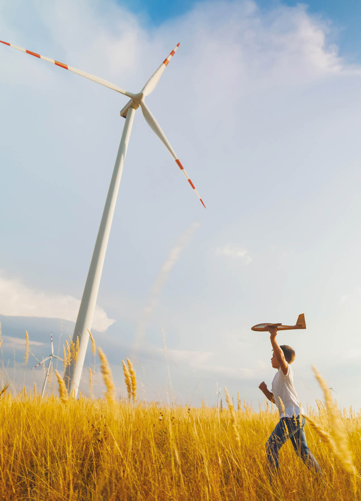 A Caucasian boy enjoys running through a field, toy airplane aloft, with towering wind turbines spinning in the background on a sunny day.